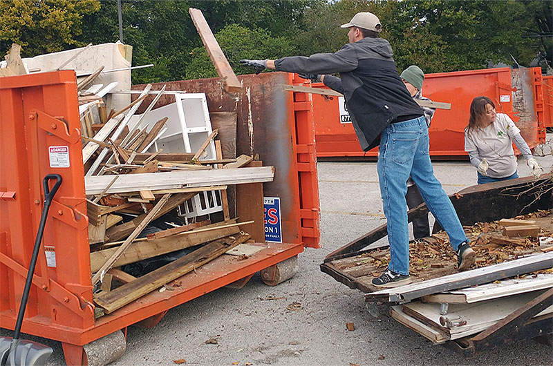 man standing on flatbed trailer throws lumber into orange dumpster