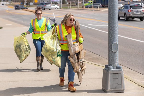 two ladies in safety vests carrying trash along a roadway