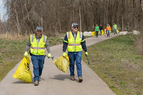 two men in yellow safety vests carrying filled trash bags out of woods on a trail